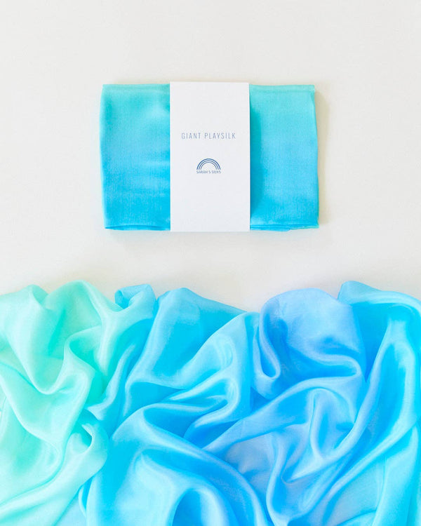 A folded turquoise Sarah's Silk Giant Sea Playsilk with a white paper band labeled "GIANT PLAY SILK" lies on a white surface. Below it, the same playsilk is unfurled, displaying a gradient of blues stretching to teal, inviting open-ended play and sparking imaginative adventures.