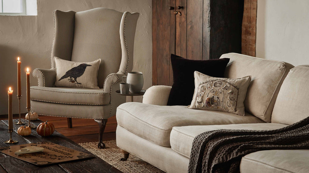 Cozy living room with striped armchair, beige sofa, Coral & Tusk Blackbird with Acorn Pillow, Halloween-themed pillows, lit candles, small pumpkins, and a Ouija board on a wooden coffee table. Warm neutral rustic decor.
