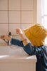 A child wearing a yellow hat and blue shirt, sporting the Mini Kyomo Watch - Cherry Bomb, stands at a white sink and reaches for the faucet as sunlight streams through the window.