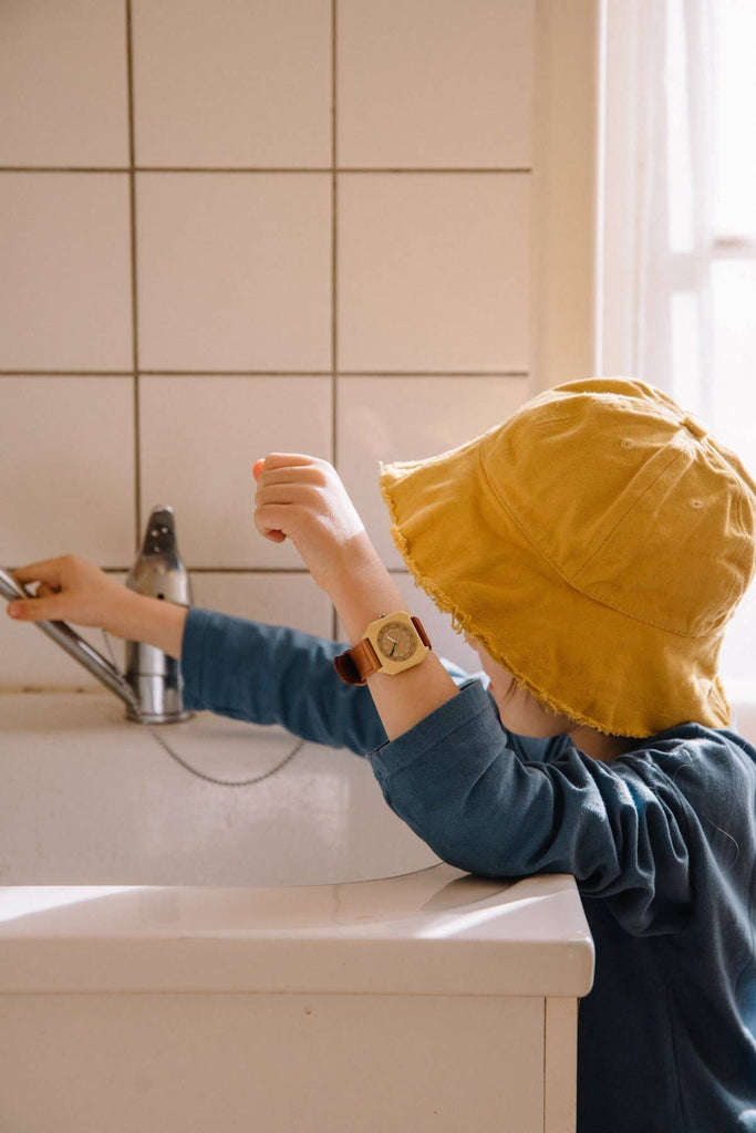 A child wearing a yellow hat and blue shirt, sporting the Mini Kyomo Watch - Cherry Bomb, stands at a white sink and reaches for the faucet as sunlight streams through the window.