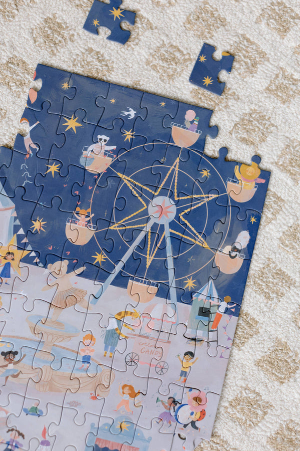 The A Carnival of Wonders 100 Piece Puzzle for ages 5+ features a whimsical carnival scene with a Ferris wheel, riders, and a crowd. Two unplaced pieces rest on a textured rug as the playful image nears completion.