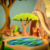 A colorful wooden diorama displays a jungle scene with three tigers. Two tigers sit beside a cave, while one stands near a small blue and green pond. The background features trees, including the Bumbu Wooden Acacia Tree Set, and rock formations, all set against a pale green backdrop.