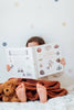 A child lies under a brown blanket, feet peeking out, reading "Our Big Book of Sports: A Collection of 100+ Sports Words." A teddy bear sits nearby, and pastel polka dots decorate the background.