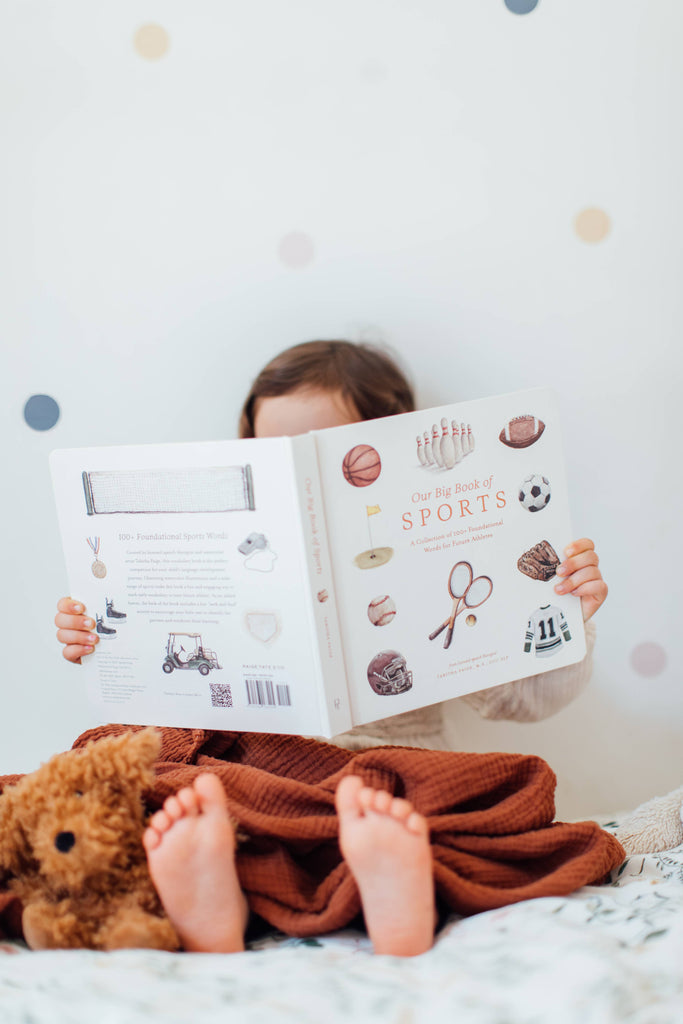 A child lies under a brown blanket, feet peeking out, reading "Our Big Book of Sports: A Collection of 100+ Sports Words." A teddy bear sits nearby, and pastel polka dots decorate the background.
