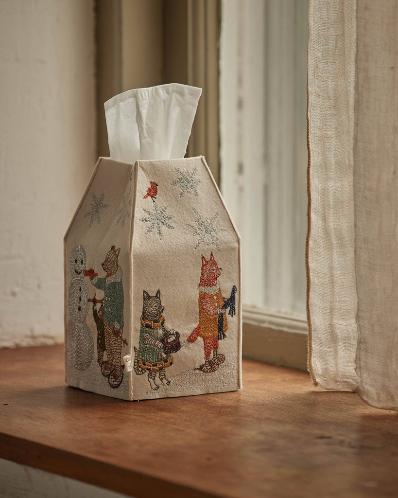 The Coral & Tusk Snow Day Tissue Box Cover, featuring embroidered cats in winter clothes and a snowman, sits on a wooden windowsill beside a sheer curtain. White tissues peek from the top—an adorable accent for snow day decor.