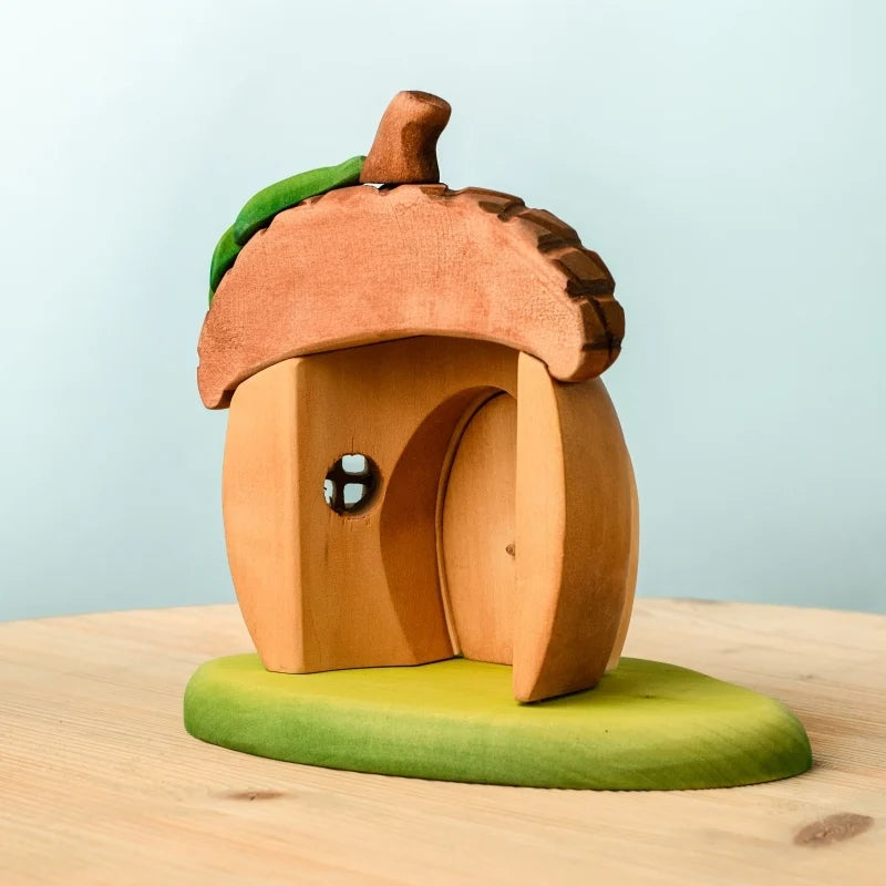 The Bumbu Handmade Wooden Acorn House, featuring a green roof and open door on a green base, sits on a light wooden surface against a pale blue background—a charming Montessori Waldorf toy.