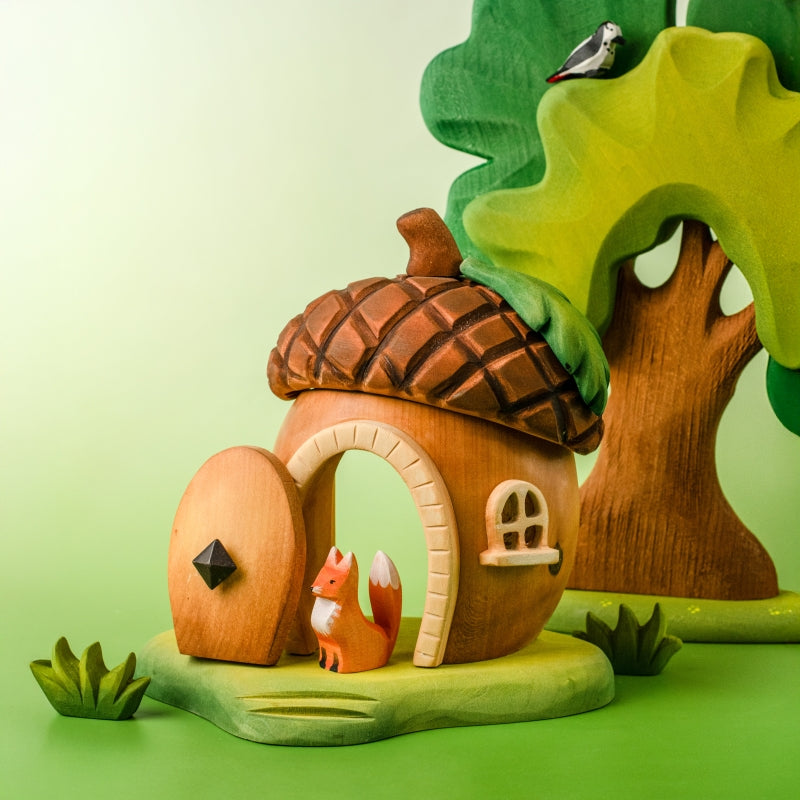 The Bumbu Handmade Wooden Acorn House is a charming fairy dwelling with an open door, a small fox at the entrance, green grass, and a tree with a bird—an ideal Montessori Waldorf toy scene.
