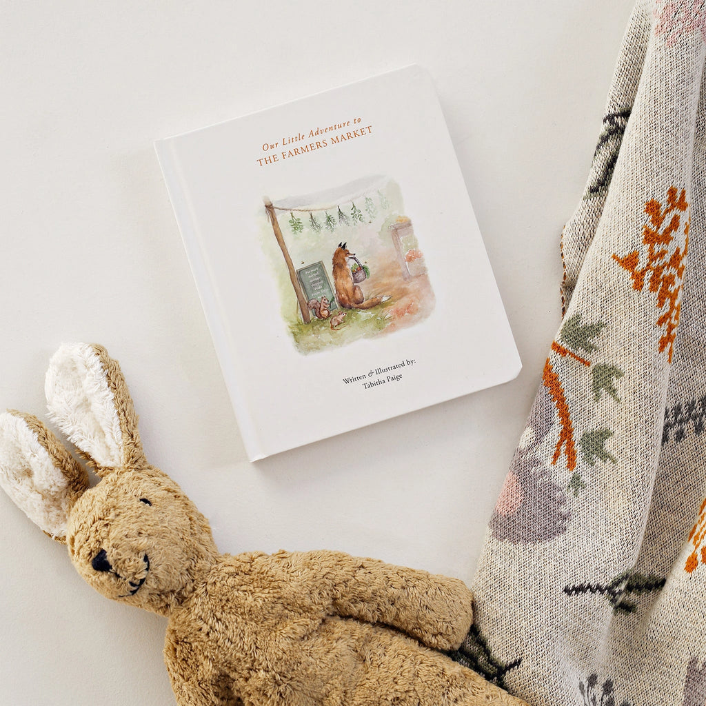 A children's book titled "Our Little Adventures To the Farmers Market - Board Book," perfect for teaching early language concepts, lies next to a beige and brown plush bunny toy and a patterned blanket. The book cover features an illustration of woodland animals at a market stand, making it an ideal baby gift.