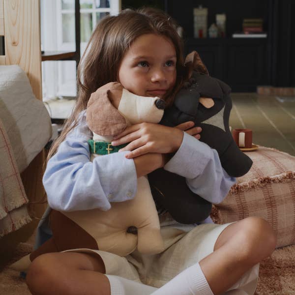 A young girl sits indoors, hugging two plush toy dogs to her chest. She looks content with natural light streaming in behind her, surrounded by cozy pillows including the Dog Cushion - Cooper, and a rug nearby.
