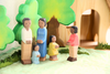 An Ostheimer Father, Cream Sweater is arranged outdoors, standing on a green surface, with two trees and a wooden structure in the background. These handcrafted wooden toys feature two adults and three children in brightly colored clothes, perfect for imaginative play.