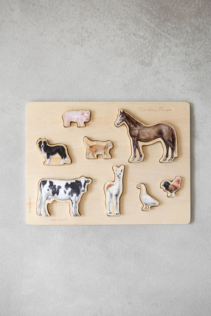 Farm Animals Wooden Puzzle: A Tabitha Paige Puzzle features a wooden puzzle board with removable animal pieces—a pig, dog, horse, cow, alpaca, duck, chicken, and cat—arranged on a light surface. This charming farm animal puzzle is an ideal toddler educational toy and classic wooden peg puzzle that encourages fine motor skills and early learning through play.