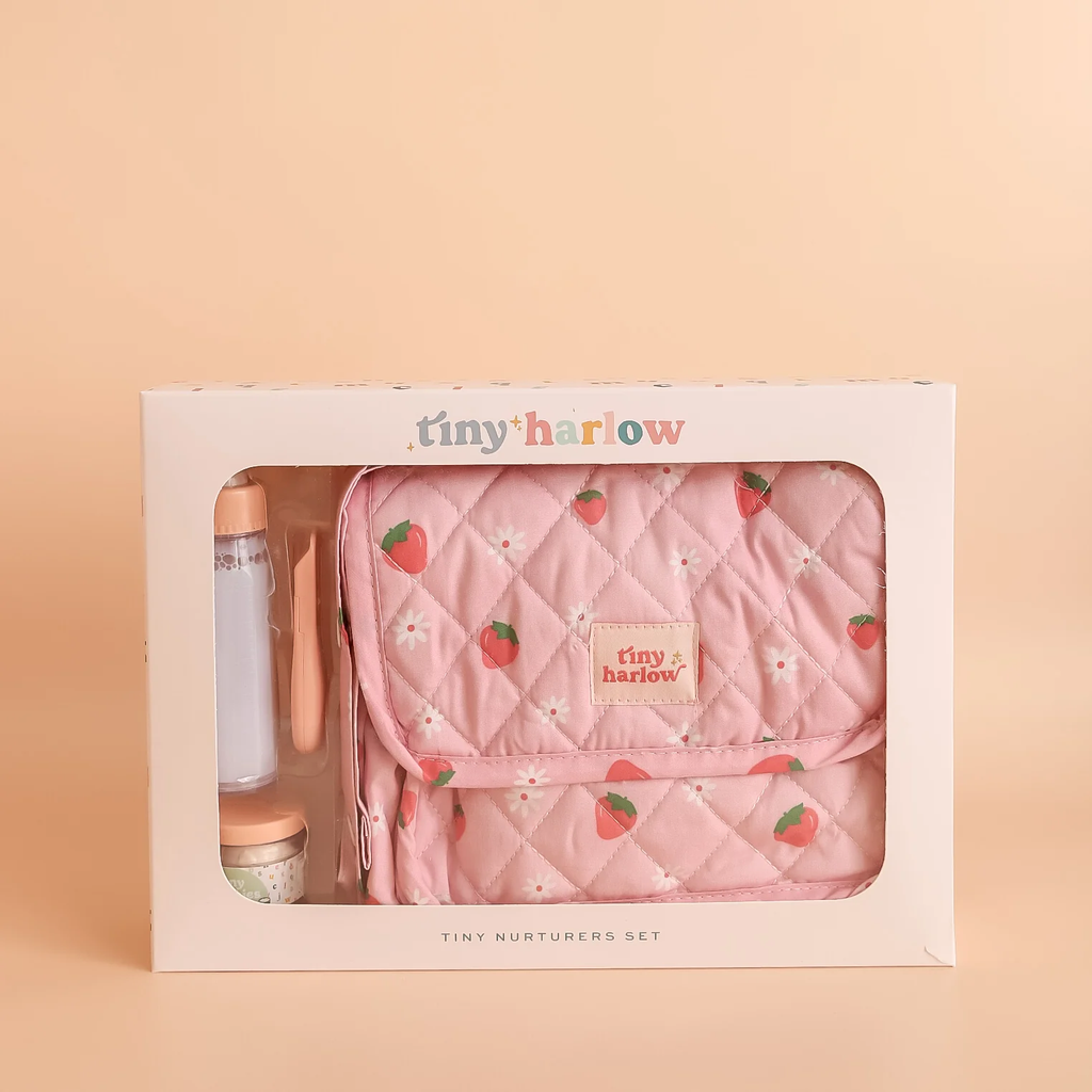 The Tiny Nurturers Set - Strawberry Picnic For Dolls comes in a box and includes a pink quilted doll nappy bag with strawberry and flower print, a toy bottle, spoon, and a magic baby doll cereal jar, all displayed against a beige background.