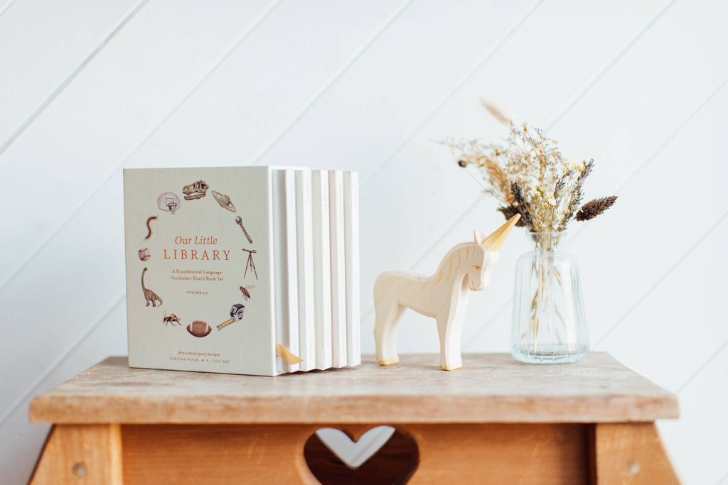 Our Little Library Vol. 3 (5 Board Books) is displayed on a wooden stool with a heart cutout, accompanied by a unicorn figurine and dried flowers—an ideal baby shower gift or tool for early language development.