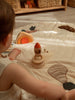 A child sits on a quilted play mat, playing imaginatively with the Sabo Concept Rocket with Bear and stackable arches, surrounded by natural wood toys displayed on shelves.