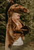A smiling child stands outdoors surrounded by green trees, wearing the Costume - Brown Horse, featuring a horse head hood and hoof-like sleeves.