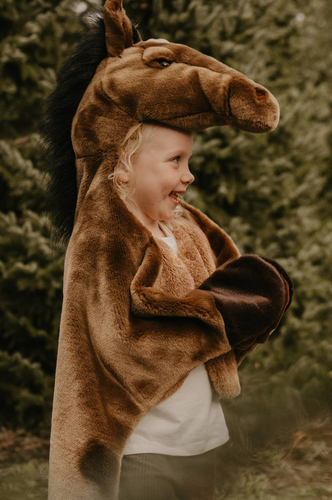 A smiling child stands outdoors surrounded by green trees, wearing the Costume - Brown Horse, featuring a horse head hood and hoof-like sleeves.