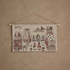 The Coral & Tusk North Pole Advent Calendar features embroidered buildings, people, stars, animals, and festive Christmas details on beige fabric, displayed against a textured light brown wall.