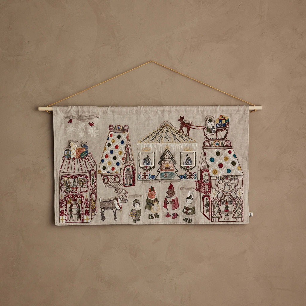 The Coral & Tusk North Pole Advent Calendar features embroidered buildings, people, stars, animals, and festive Christmas details on beige fabric, displayed against a textured light brown wall.