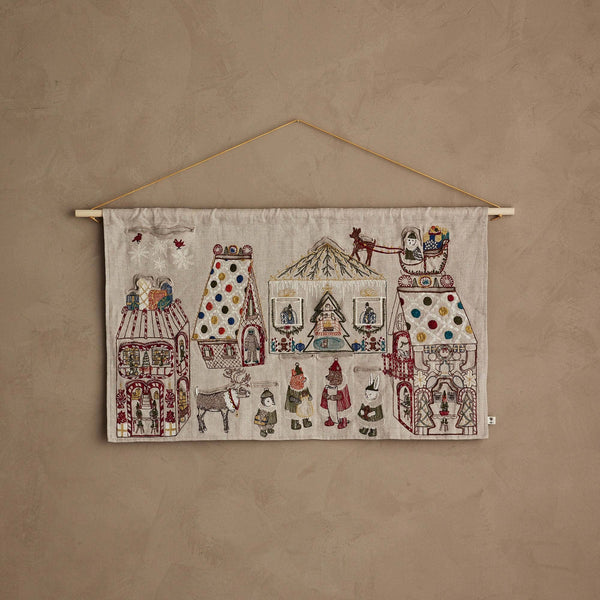 The Coral & Tusk North Pole Advent Calendar features embroidered buildings, people, stars, animals, and festive Christmas details on beige fabric, displayed against a textured light brown wall.
