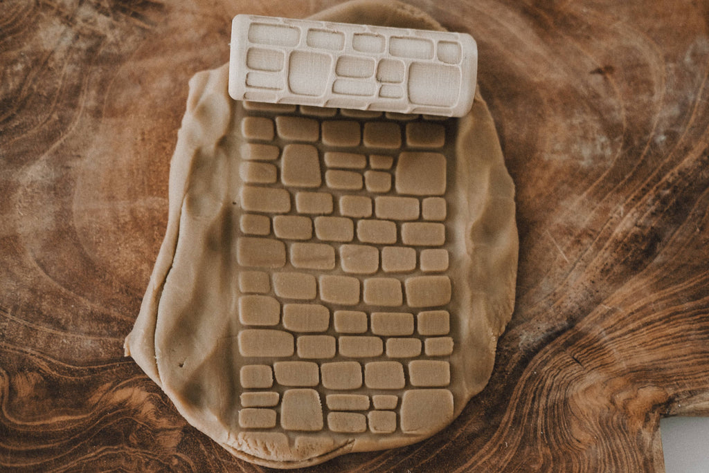The Cobblestone Path Roller - Pattern Playdough Roller is ideal for creative sensory play, as it imprints a brick pattern onto a sheet of light brown dough placed on a wooden surface.