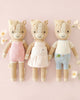 Three Cuddle + Kind Liam the Pony dolls, handmade in Peru, stand side by side in pastel pink overalls, a light pink dress, and blue shorts against a pastel pink background with flowers.