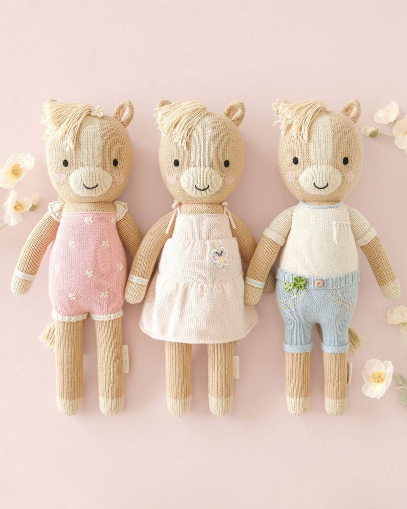 Three Cuddle + Kind Liam the Pony dolls, handmade in Peru, stand side by side in pastel pink overalls, a light pink dress, and blue shorts against a pastel pink background with flowers.