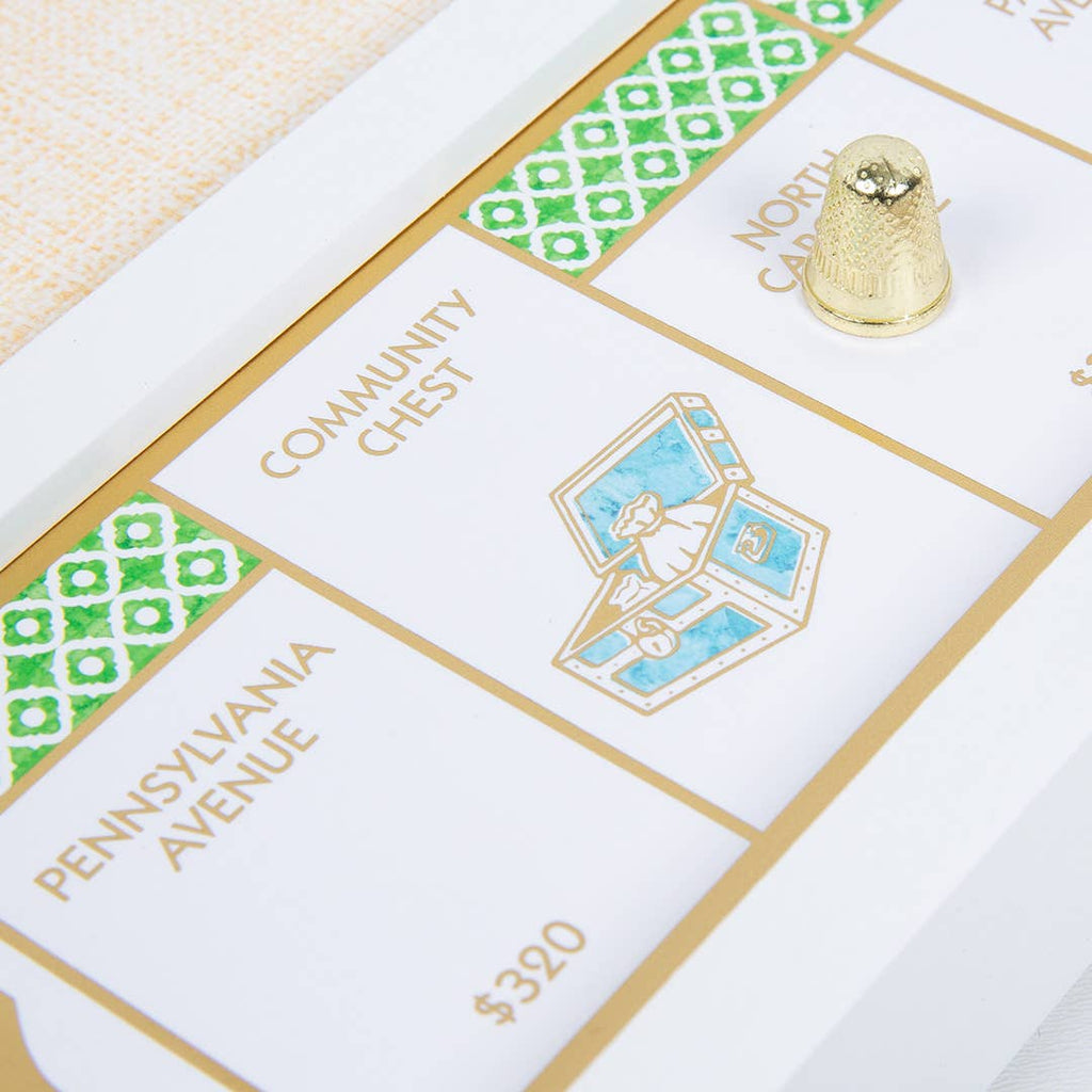 A corner of the Monopoly Bianco Edition gold foil-stamped game board featuring Pennsylvania and North Carolina Avenue properties, a Community Chest space with a treasure chest illustration, and a gold-colored thimble playing piece.