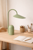 A minimalist wooden desk features the Ferm Living Arum Portable Lamp in Tea Green, an open notebook with a pencil, a basket of colored pencils, paintbrushes, and neatly arranged wooden toys on the surface.