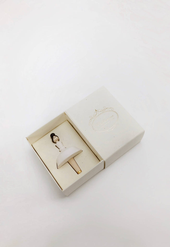 A small white box opens to reveal the delicate Wooden Ballerina, a hand-painted figurine featuring a white dress and dark hair. The elegant design includes cursive writing on the lid, and the plain white background emphasizes the simplicity and grace of its packaging.