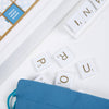 The corner of a Scrabble Bianco Edition board displays a Triple Word Score square, a row of letter tiles, and loose tiles scattered near a blue fabric drawstring bag.