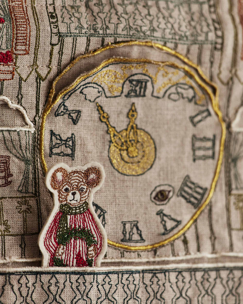 The Coral & Tusk Christmas Clock Advent Calendar features a bear in a red and green sweater in front of a large clock with Roman numerals and gold hands, all embroidered with intricate threadwork.