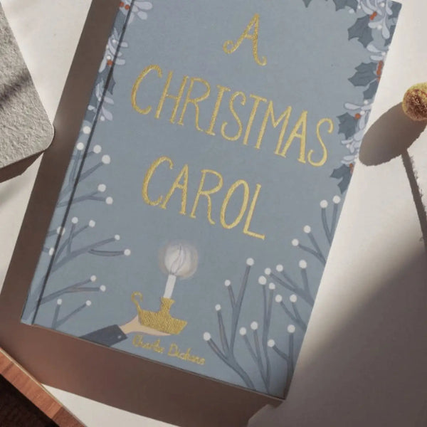 The "A Christmas Carol | Collector's Edition| Hardcover Book" rests on a white surface in sunlight, its blue cover adorned with gold lettering, holly accents, and a candle—evoking Ebenezer Scrooge’s classic tale.