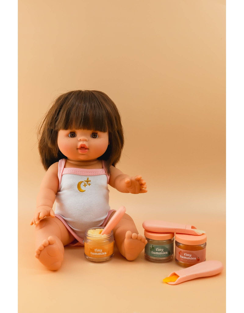 A doll with brown hair is seated against a beige background, dressed in a white outfit featuring a moon and star. Nearby are three small jars filled with colorful substances, labeled "Tiny Tummies - Apple Jelly Food," alongside two pink magic spoons for use when the doll "eats.