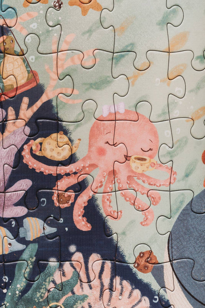 The Tales from the Deep 100 Piece Puzzle shows a pink octopus with a bow holding a teacup underwater, surrounded by coral, fish, and a yellow teapot. Made from recycled materials and perfect for ages 5+.