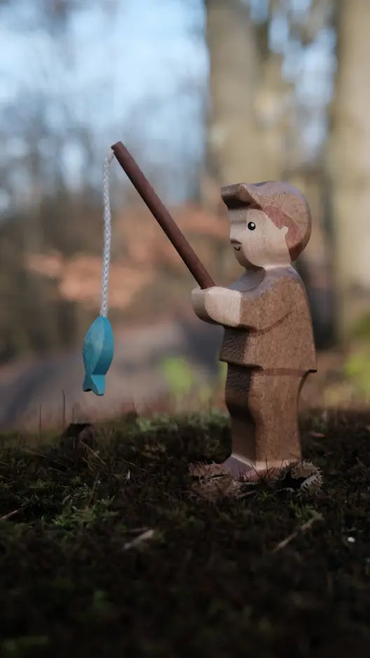 The Handmade Holzwald Fisherman, a wooden child figure, stands on moss holding a fishing rod with a blue fish. Blurred outdoor trees and natural light in the background make it ideal for fans of nature-inspired toys.