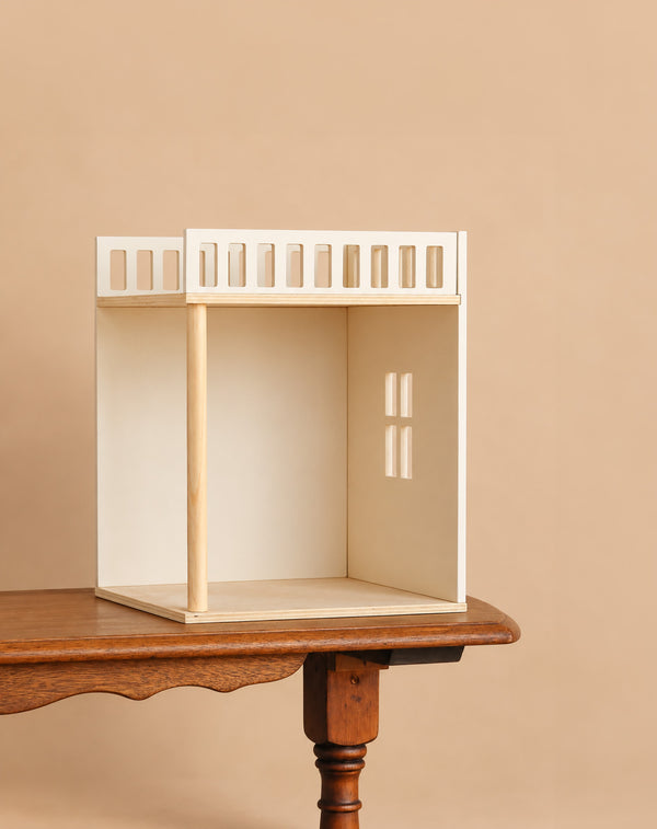 A minimalist, unfinished wooden dollhouse rests on a wooden table against a beige background, featuring one visible window with sunlight streaming through—a perfect setting to add the Maileg House of Miniature Bonus Room - Bedroom and create a cozy retreat inside.