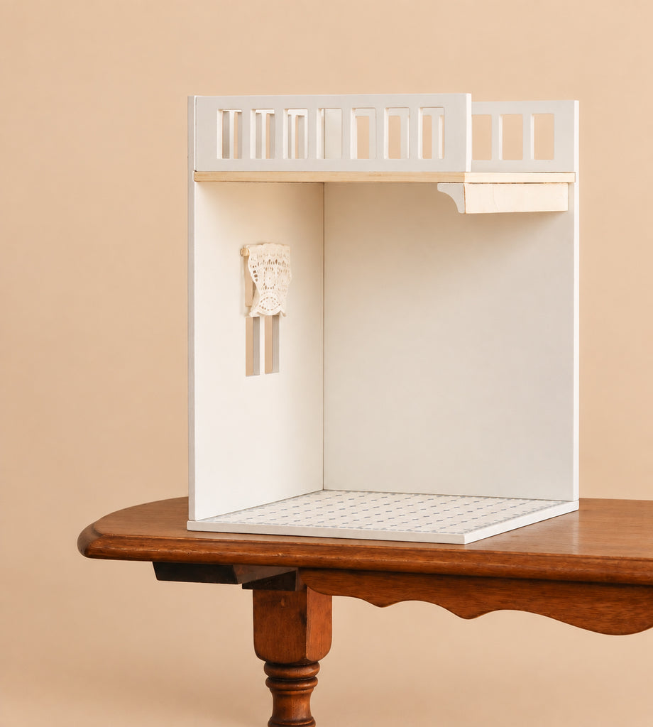 A small, white Maileg House of Miniature Bonus Room - Bathroom with cutout windows, a decorative wall piece, and a lofted bed sits on a wooden table against a beige background. This open dollhouse bathroom can serve as the perfect bonus room or be complemented with a charming wooden dollhouse accessory.