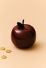 The Ferm Living Apple Money Bank, in glossy dark red with a coin slot on top, sits on a light beige surface beside three gold coins—a stylish and charming way to save your change.