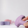 A hand holds a purple, U-shaped Grimm's Casa Coral Building Block made of handpainted wood with white details. In the background, two more wooden pieces—one pink, one purple—rest on a white surface, ideal for toddlers.