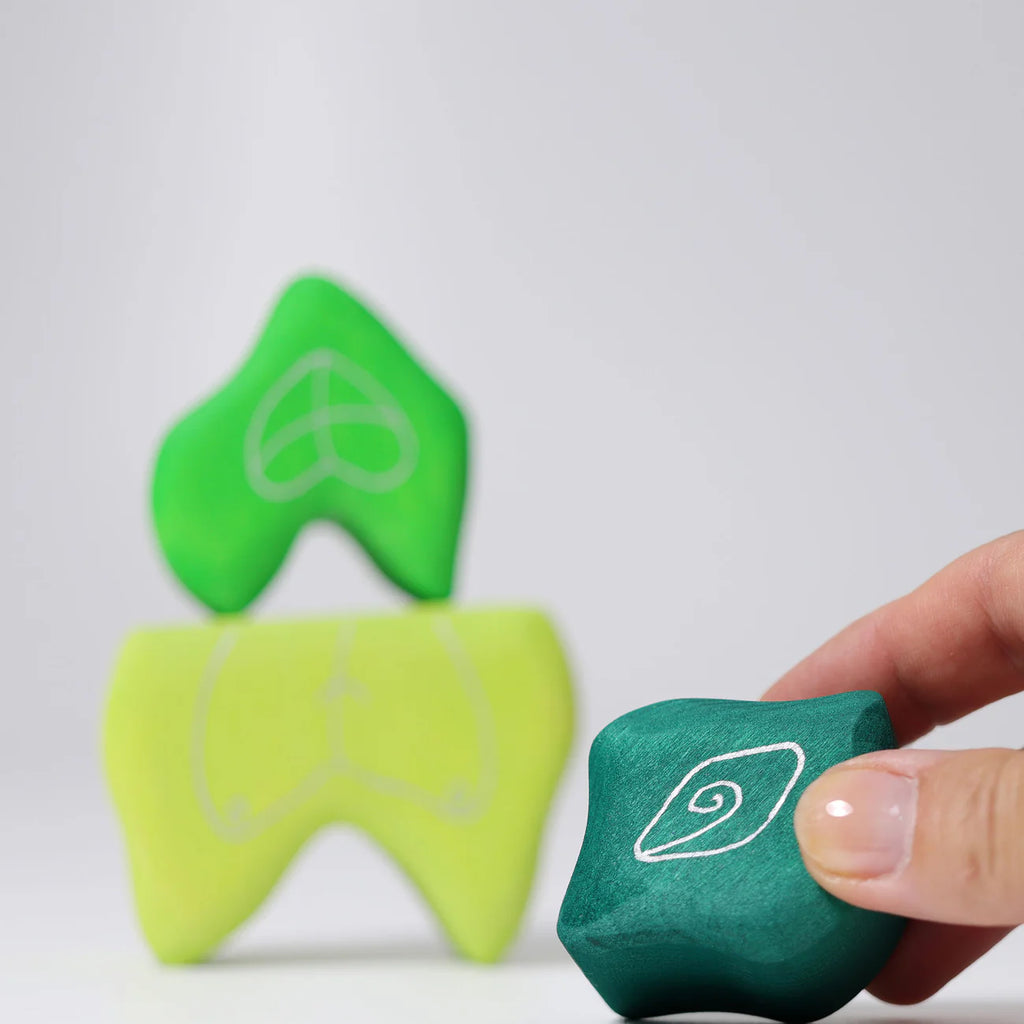 A hand holds a green Grimm's Tower Leaves Building Block with a white spiral, near two stacked blocks in green and yellow featuring white line patterns, all set against a plain light background.