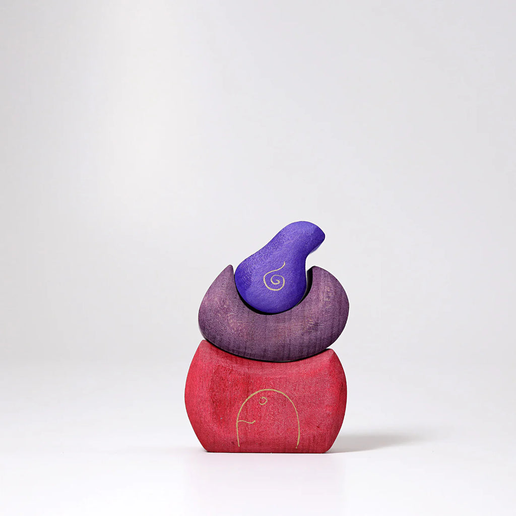 Grimm's Small Bakery Building Blocks feature three organic pieces in red, purple, and violet, stacked in a minimalist bird shape. Smooth surfaces and gold swirl details make them perfect for imaginative wooden play against a white background.