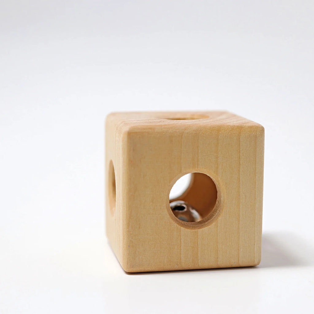 The Grimm’s Cube with Bell (ships in 2-3 weeks), a lightweight and compact wooden toy featuring smooth edges and circular holes on each side, rests on a white surface. Through one of the openings, a metal bell can be seen inside the cube, making this a delightful non-toxic baby toy.