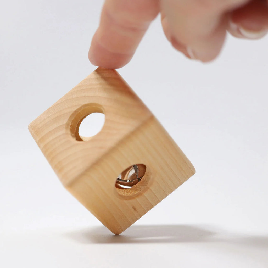 A hand balances Grimm’s Cube with Bell—a handcrafted wooden toy cube with circular holes—on its edge against a white background.