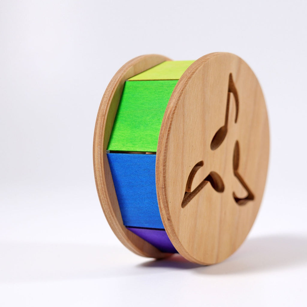 Grimm's Sound and Color Wheel is a non-toxic wooden baby motor skill toy with a cylindrical shape, green, blue, and purple segments, musical note cutouts, and stands upright on a white background.