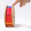 A hand points to Grimm's Sound and Color Wheel, a non-toxic wooden toy drum with purple, red, and orange sections on a white background—ideal for developing baby motor skills.