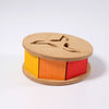 Grimm's Sound and Color Wheel is a non-toxic wooden toy with a round base and top, featuring three colorful sides—yellow, orange, and red—and three cutouts on top to help develop your baby’s motor skills.