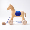 Grimm's Horse Holly is a wooden pull-along horse featuring a blue cloth saddle, four wheels, and a rope with a snail shell. Its minimalist design makes it a charming toddler toy, pictured against a plain white background.