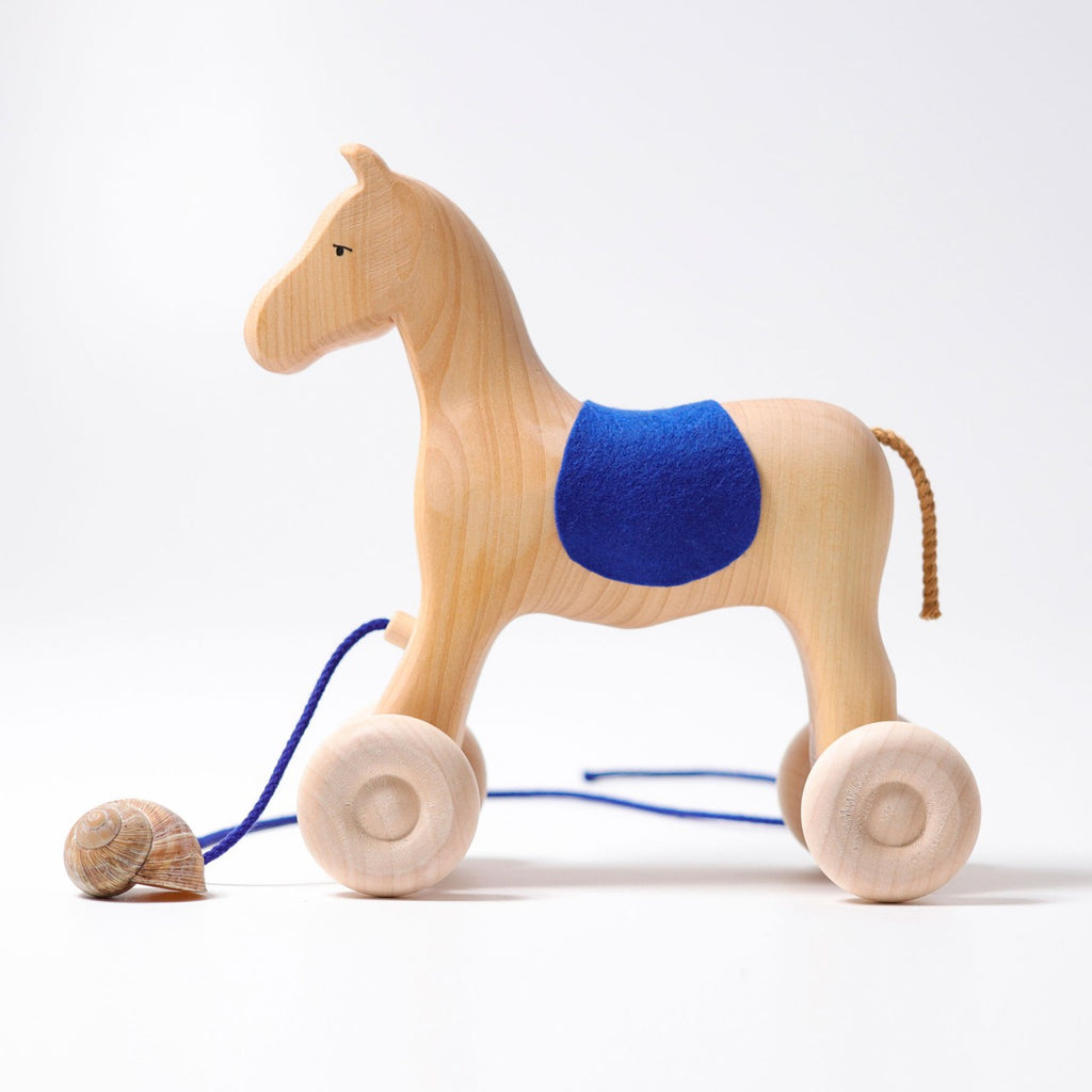 Grimm's Horse Holly is a wooden pull-along horse featuring a blue cloth saddle, four wheels, and a rope with a snail shell. Its minimalist design makes it a charming toddler toy, pictured against a plain white background.