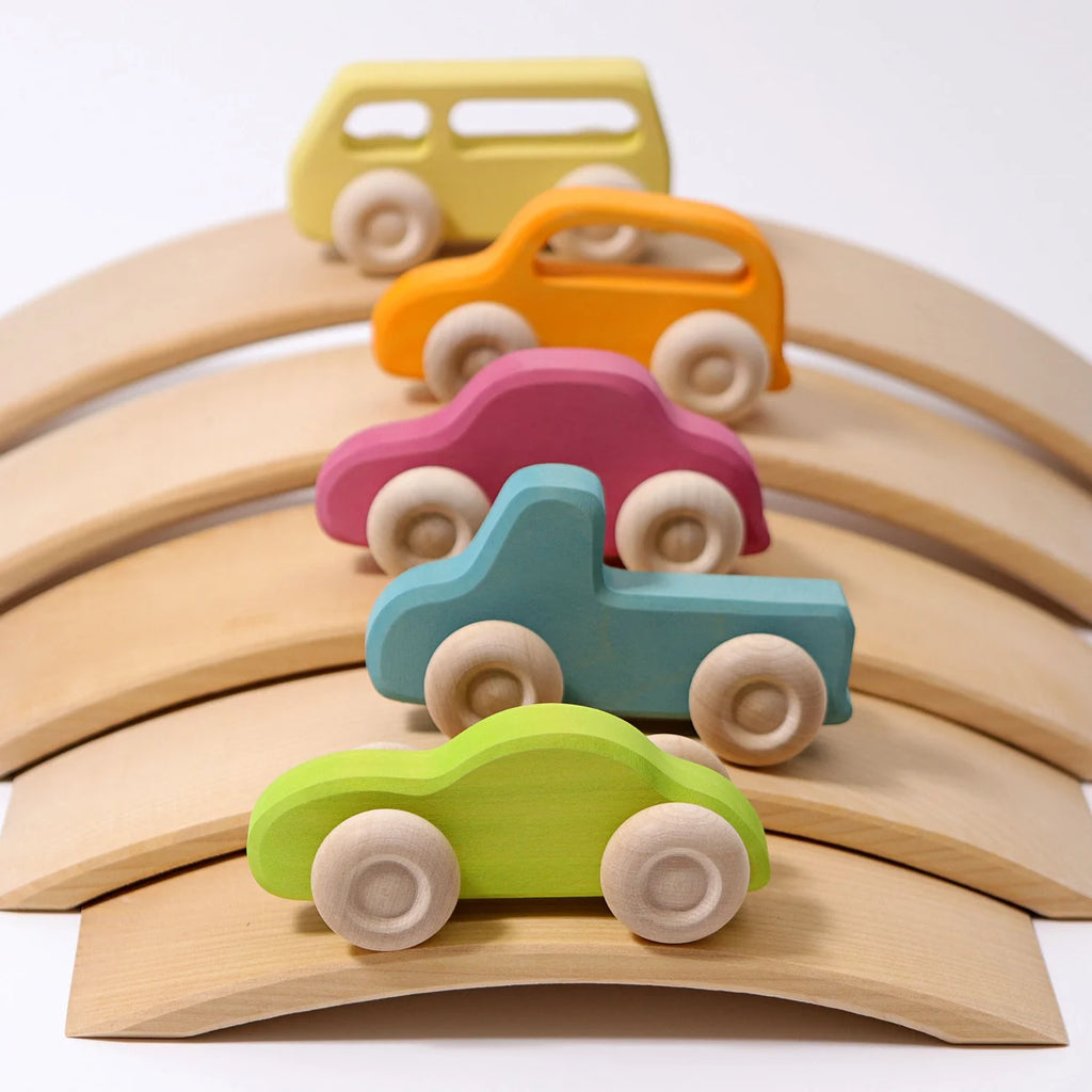 Five colorful Grimm’s Wooden Cars Slimline, finished with natural oils and each in a different vibrant shade—green, blue, pink, orange, and yellow—are lined up on curved wooden ramps against a white background; perfect as eye-catching toddler toys.