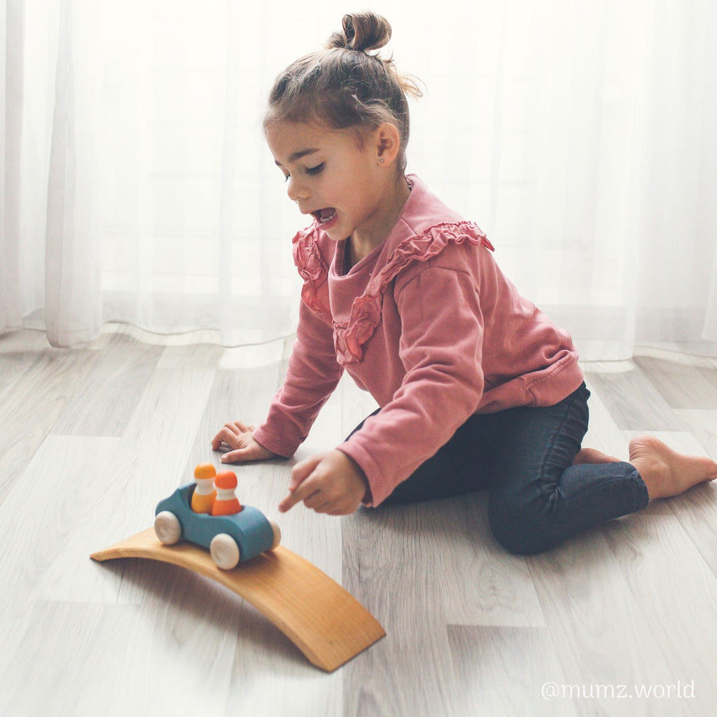 A young child with a top knot hairstyle, wearing a pink sweater and black pants, is sitting on the floor playing with a Grimm's Wooden Blue Convertible Car featuring a non-toxic color stain on a curved wooden ramp. The backdrop is a sheer white curtain, creating a bright and airy atmosphere.